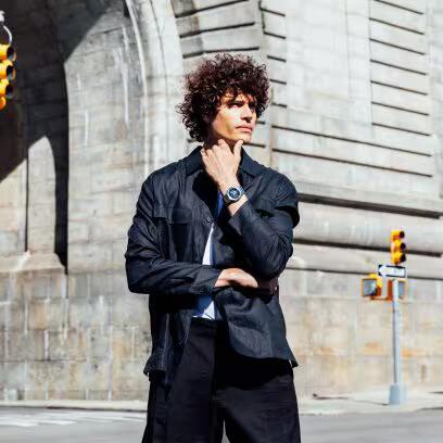 Man wearing CASIO G-SHOCK Mod. OAK G-STEEL PETROL BLUE watch stands outside urban backdrop.