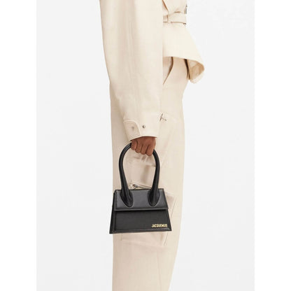 Side view of Jacquemus Le Chiquito Moyen Black Bag with gold logo, held by model in beige outfit, featuring handle and magnetic flap closure.
